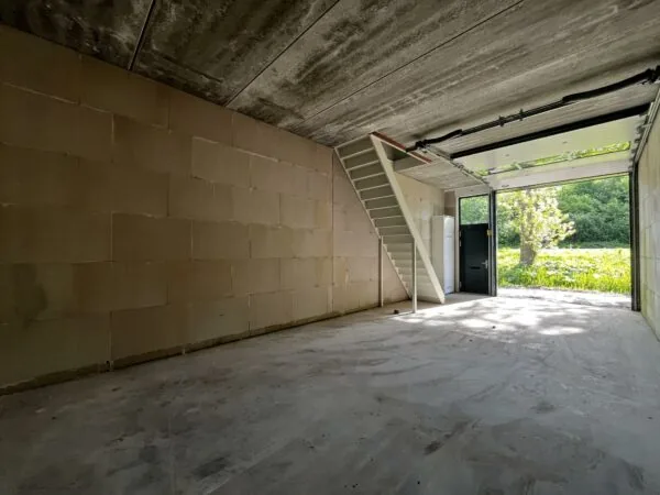 Interieur van een lege garage met trap en open garagedeur aan Broekdijk West 19-4, uitkijkend op een groene omgeving.
